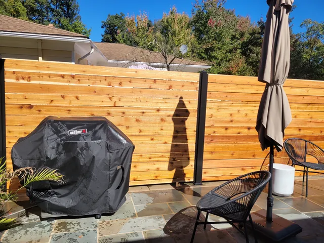 Horizontal cedar privacy panels with black metal posts behind a stone patio with a covered grill, umbrella and chairs