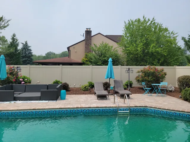 Tan vinyl privacy fence enclosing an in-ground pool with a paver deck, lounge chairs and teal umbrellas