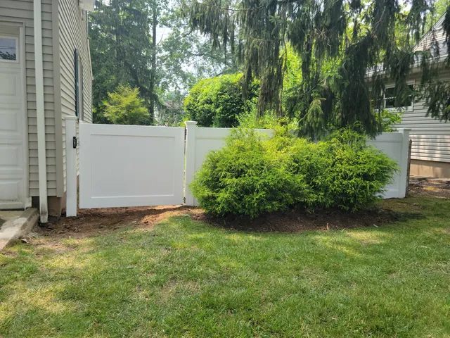 White vinyl privacy fence with a walk gate beside a house, a round evergreen shrub and lawn in front