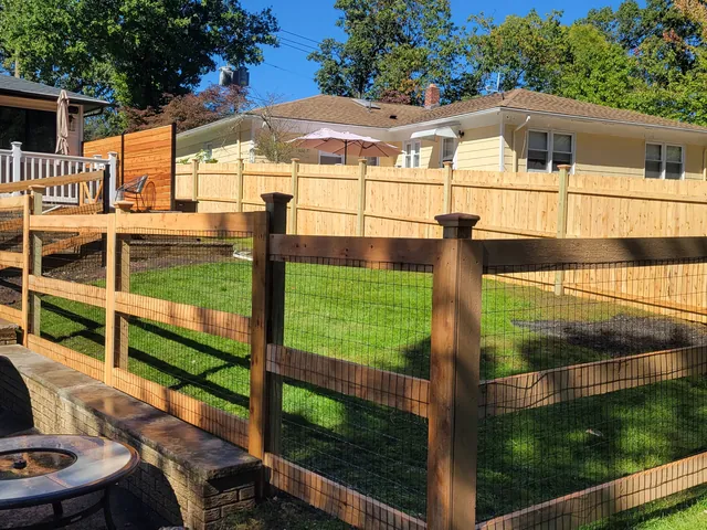 Wood three-rail fence with black wire mesh around a lawn, beside a stone patio with a fire pit and a wood privacy fence behind