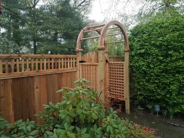 Cedar board fence with a lattice top and matching gate under an arched wood arbor, framed by shrubs