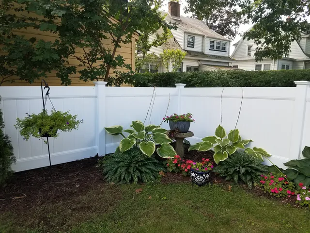 White vinyl privacy fence behind a garden bed of hostas, red flowers and a hanging planter