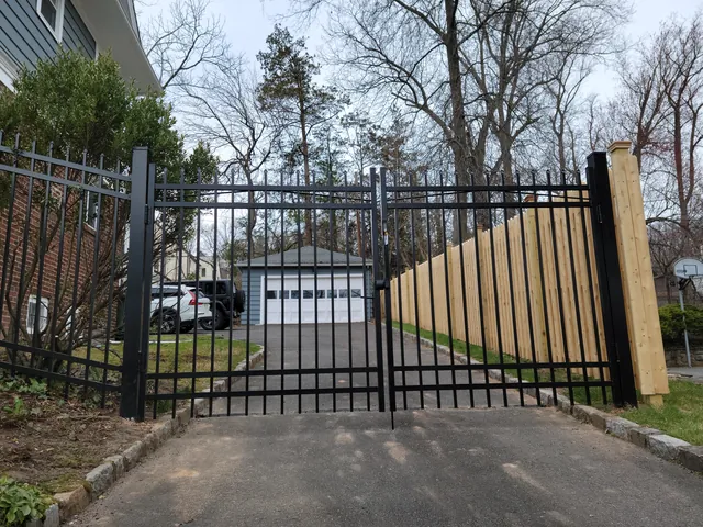 Black aluminum double driveway gate beside a tall wood privacy fence, with a detached garage at the end of the driveway