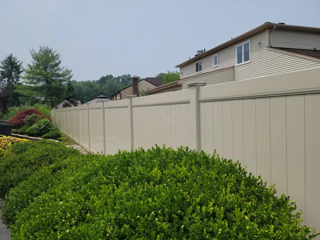 Tan vinyl privacy fence running behind clipped shrubs beside a two-story house