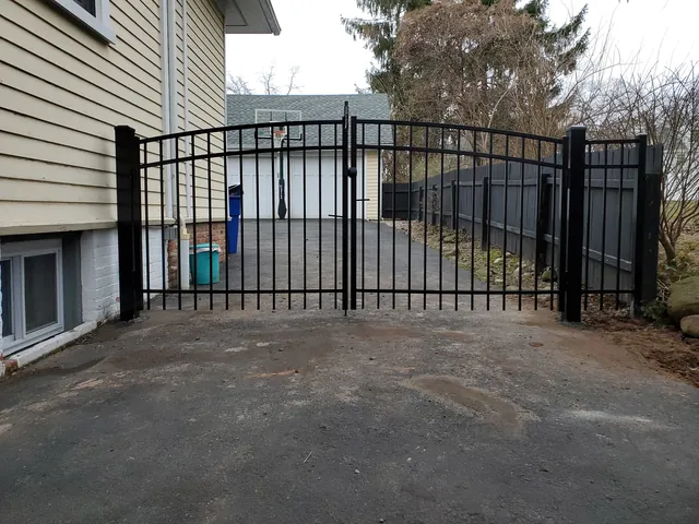 Black aluminum double driveway gate with an arched top between a house and a dark board privacy fence